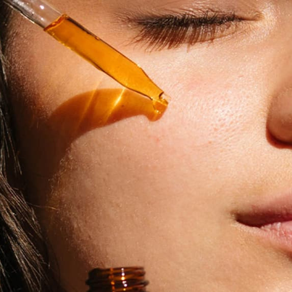 Person applying a dropper of oil to their hair and face with a bottle of oil in hand.